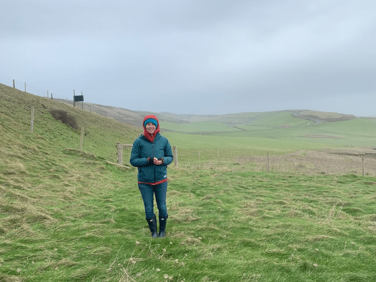 full body shot of Robin standing on an expansive green field bundled up under a gray ominous sky.