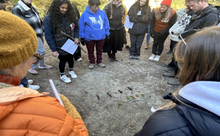 A group of people standing in a circle looking at a sculpture made up of sticks on the ground.
