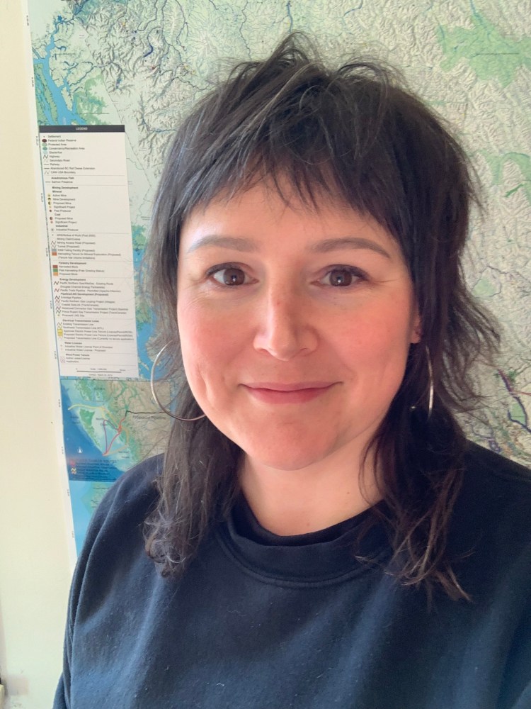 Headshot of Estraven in front of a map, they have dark hair and are wearing a black shirt