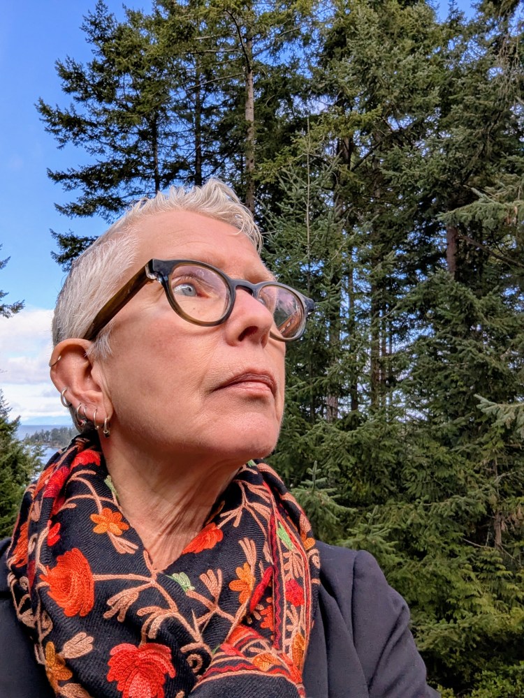 Headshot of Cate Sandilands in front of tall pine trees looking forwards the sky. Cate is wearing a colorful black and red scarf and black glasses