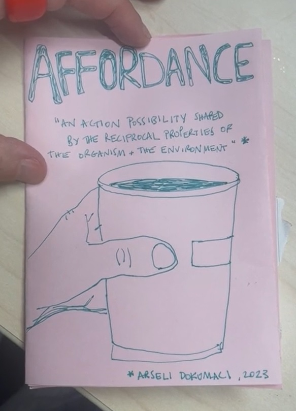 A hand holds a pink zine page. On the page is a sketch of a hand holding a paper coffee cup with the text “Affordance: ‘An action possibility shared by the reciprocal properties of the organism + the environment’. Arseli Dokumaci, 2023.”