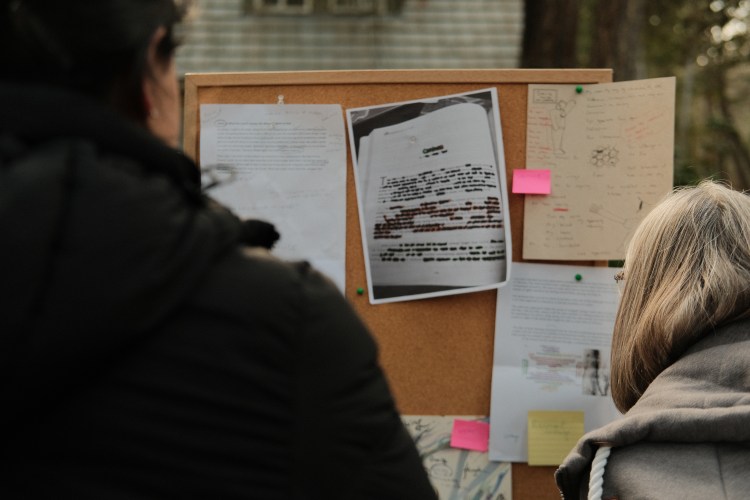 A person dressed in black stands in the foreground. In front of them is a bulletin board pinned with typed stories, drawings, and sketches. Sticky notes rest on these stories with people’s thoughts.