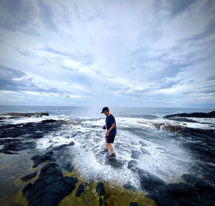 Sue standing in the muddle of a tidepool with the tide flowing in a white rush and gray clouds overhead