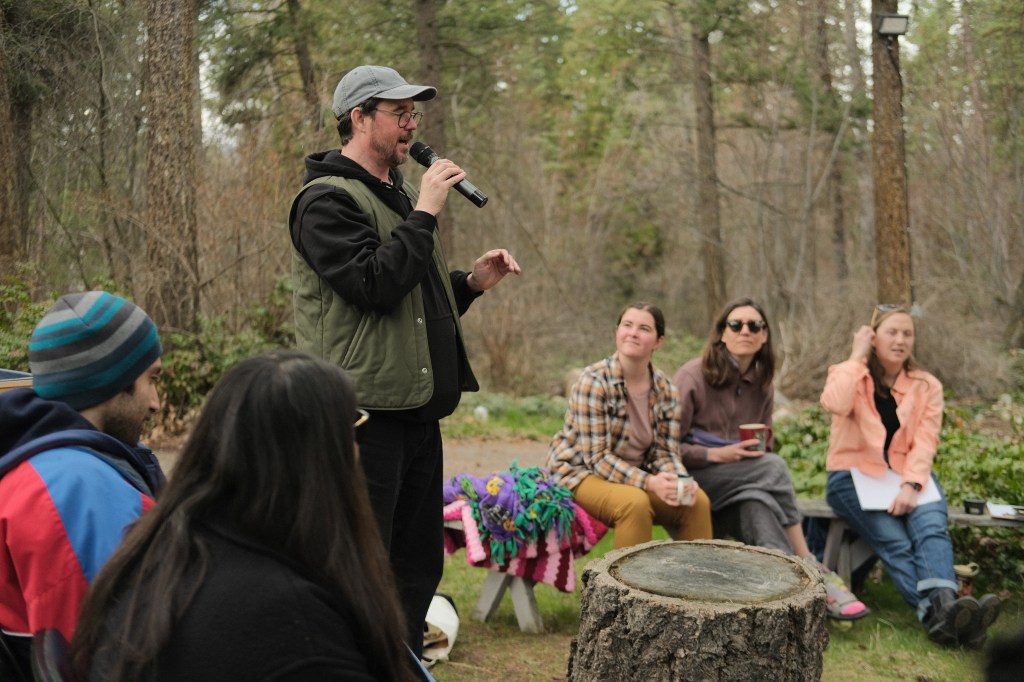 A man in a black sweater and green vest stands outside, holding a microphone. He speaks to people sitting on benches, holding mugs of tea.