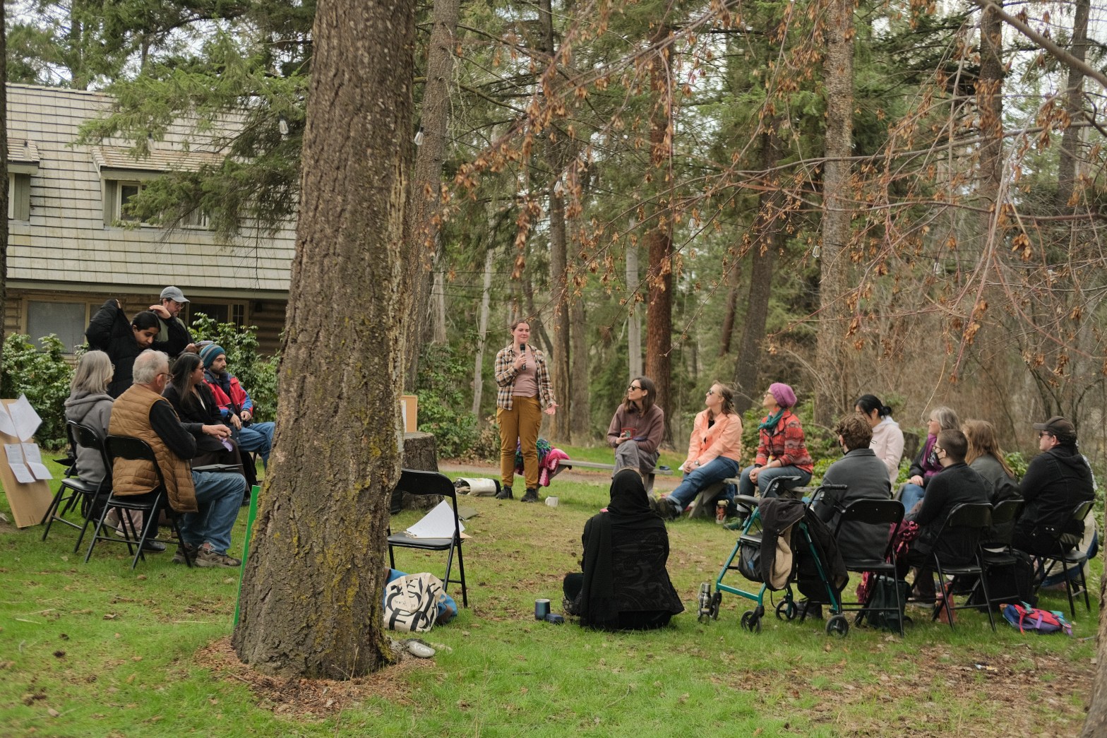 People sit on black folding chairs in a circle, outside on the vibrant grass with a cottage-looking building behind them. Most are dressed in light coats and sweaters against the early spring chill.