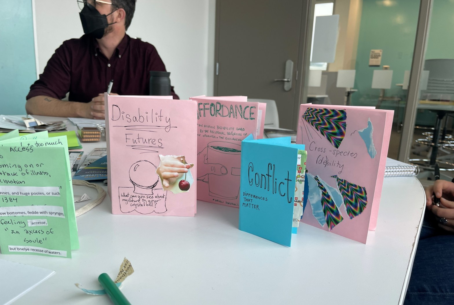 Five zines stand upright on a white table, made from sketches, collage, and text, they are colourful. Their titles read : “Access, Disability Futures, Affordance, Conflict, and Cross-species (de)bility”. A person in a maroon shirt sits behind the zines and wears a black mask. A pair of white hands rest in the right-side of the image.