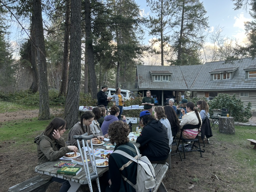 A large group of people seated around a wooden table in front of the FEELed Lab at sunset
