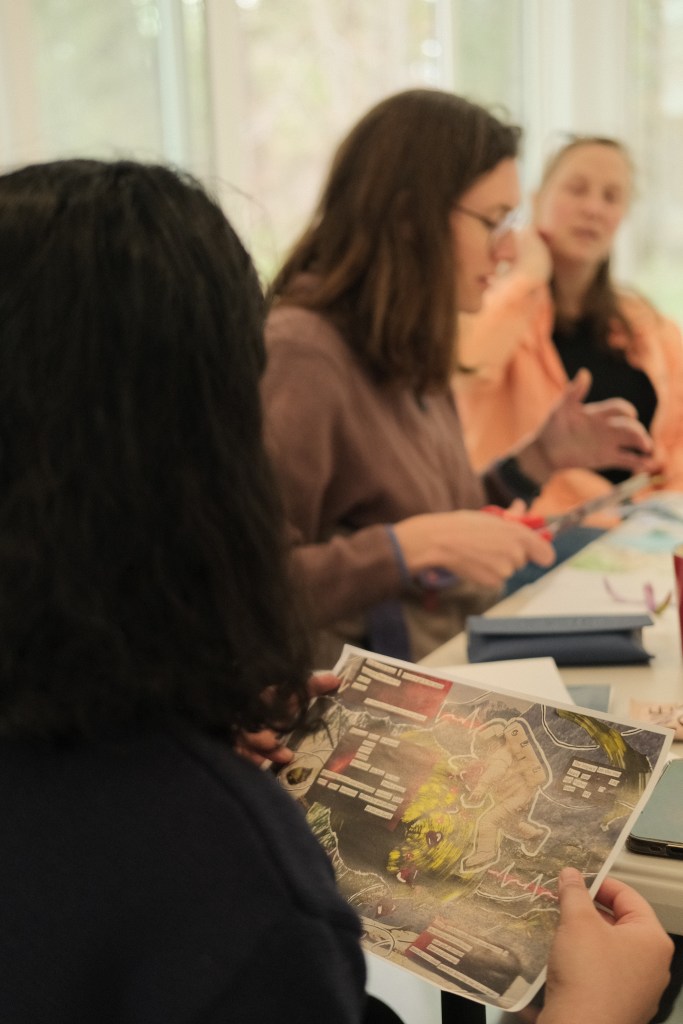 The back of a person with black hair and a black sweater is visible. They hold a piece of paper with a collage bursting with colour, reds, greens, whites, greys. An astronaut in a white suit is positioned on the edge of a swirling vortex of green. There is blurred cut out text on the page, and a painted white line representing an EKG or heartbeat. Two other people are in the background, blurred. One is cutting a piece of paper while talking as the other seems to listen to what they are saying.