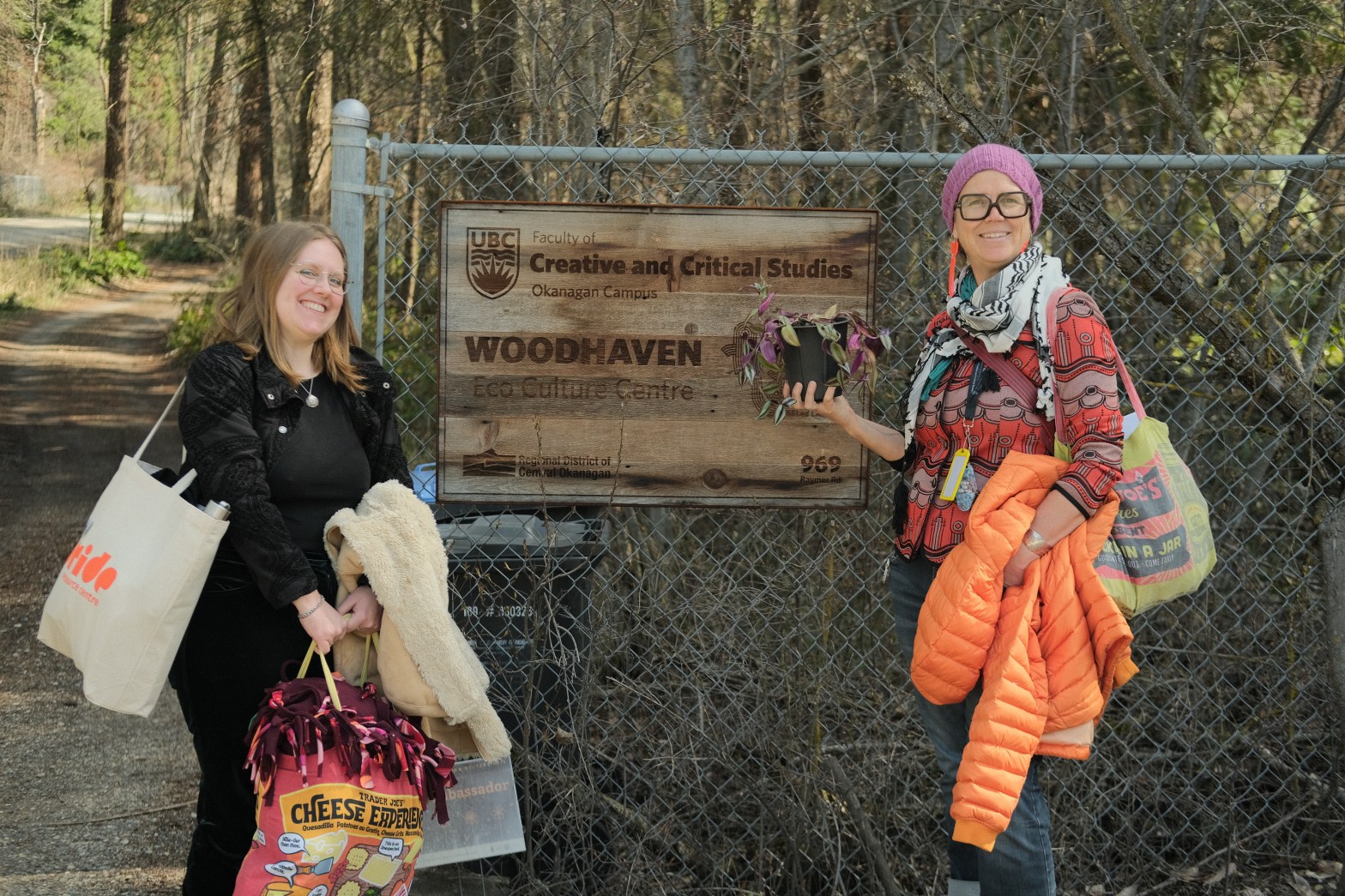 Two people pose in front of a chain-link fence, smiling brightly. A wooden sign is carved with the name “Woodhaven Eco Culture Centre”. To the right of the sign, Astrida carried an orange coat and a large bag. They hold a purple and green plant and wear brightly patterned clothes. J stands to the left, carrying a couple tote bags and a white coat. Dey wear black clothes and a seashell necklace. A gravel path to the left of J leads into the forest, trees stand to the right behind the fence.