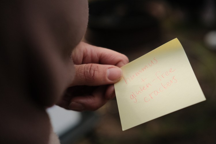 A white hand holds a yellow sticky note that says “hummus + gluten-free crackers” in red ink. The background is a blurred green and black.