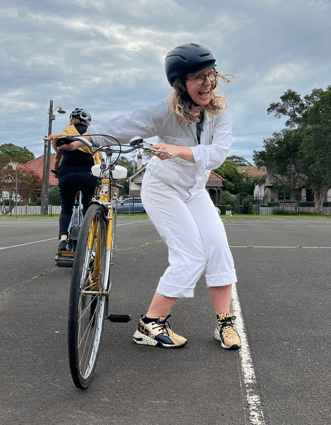 Suzie smiling down at the camera next to a yellow bicycle in a parking lot surrounded by trees