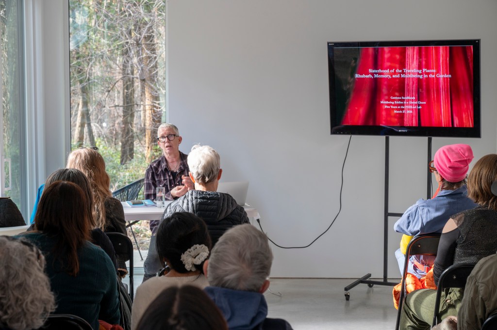 Cate Sandilands giving a talk in Woodhaven studio, the screen behind her shows a closeup of rhubarb stalks