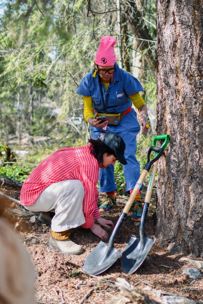Digging up the poetry book, Norah crouching on the found, Astrida leaning close in, two shovels leaning against a tree