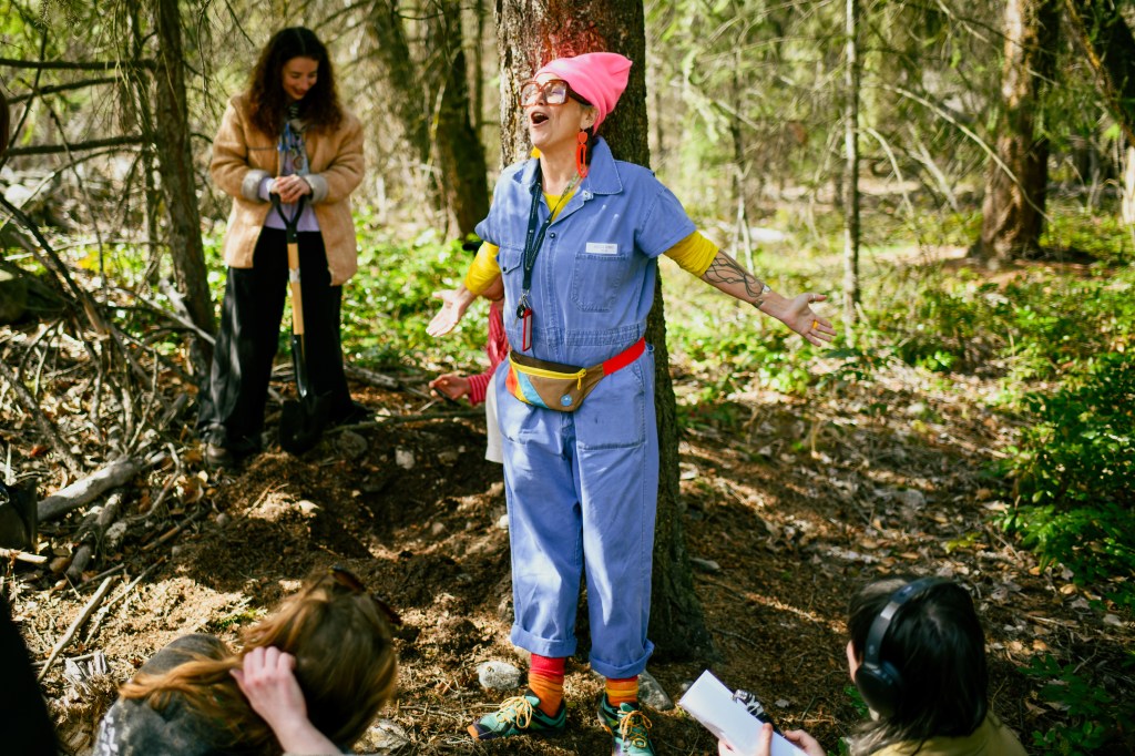 Astrida speaking expressively in front of a large tree with their arms outstretched
