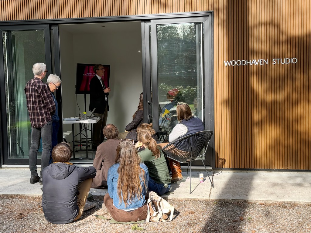 the view from outside Woodhaven studio during introductions, some people sitting on chairs and the ground outside the open sliding doors of the studio
