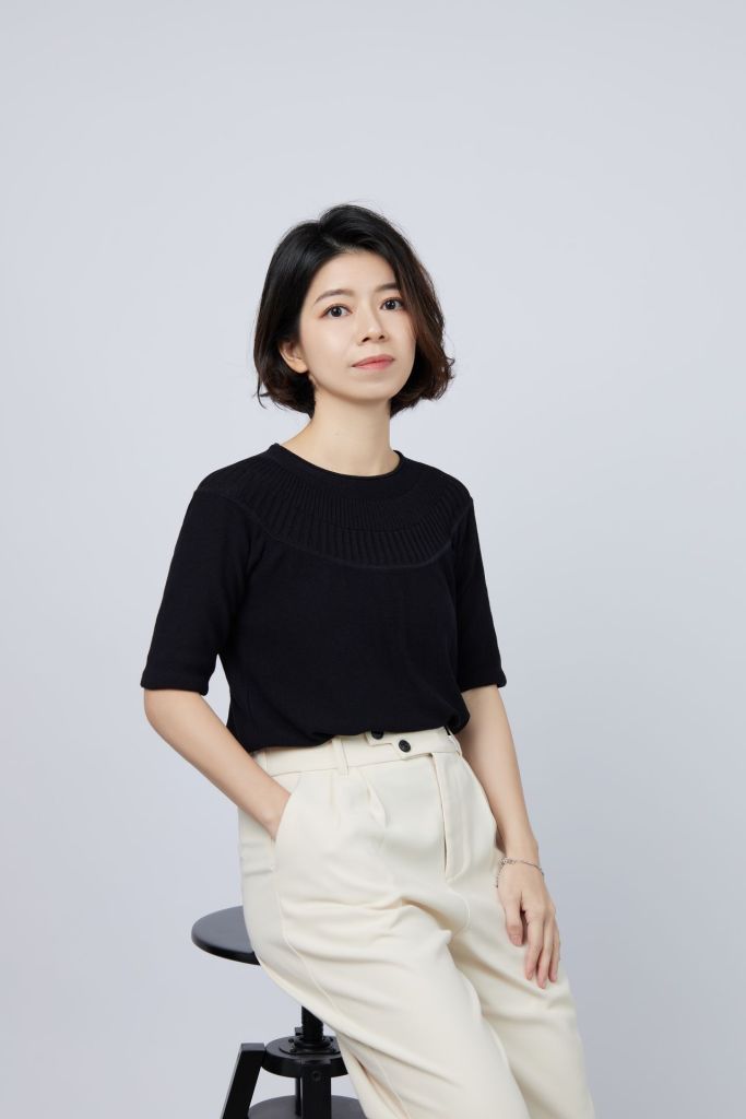Portrait of Fei leaning on a chair and looking into the camera. She is wearing beige pants and a black shirt.