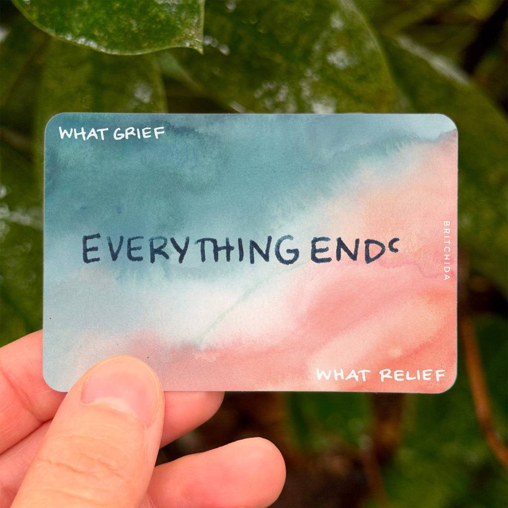 close up of a hand holding a sticker that says "Everything Ends" in the center with "What Grief" on one side and "What Relief" on the other. The sticker is ombre shading from gray/blue to red/pink.