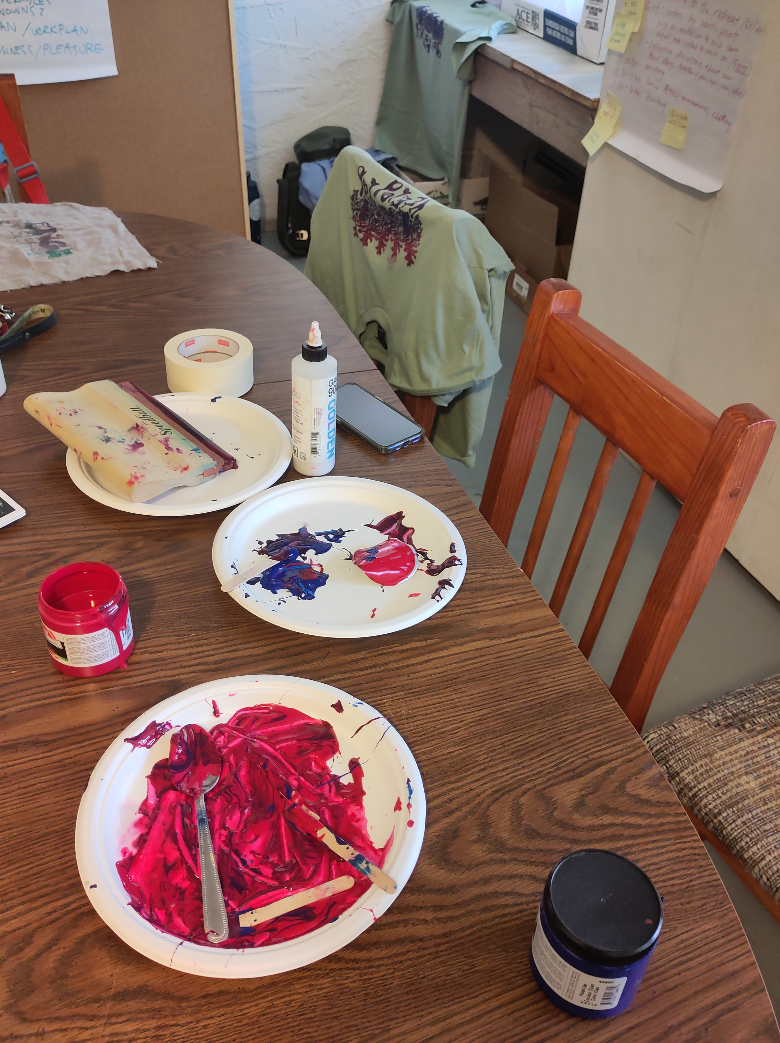Aftermath of silk screen printing on the FEELed Lab kitchen table with light green shirts hanging in the background drying. Lots of dark red and blue paint on the table