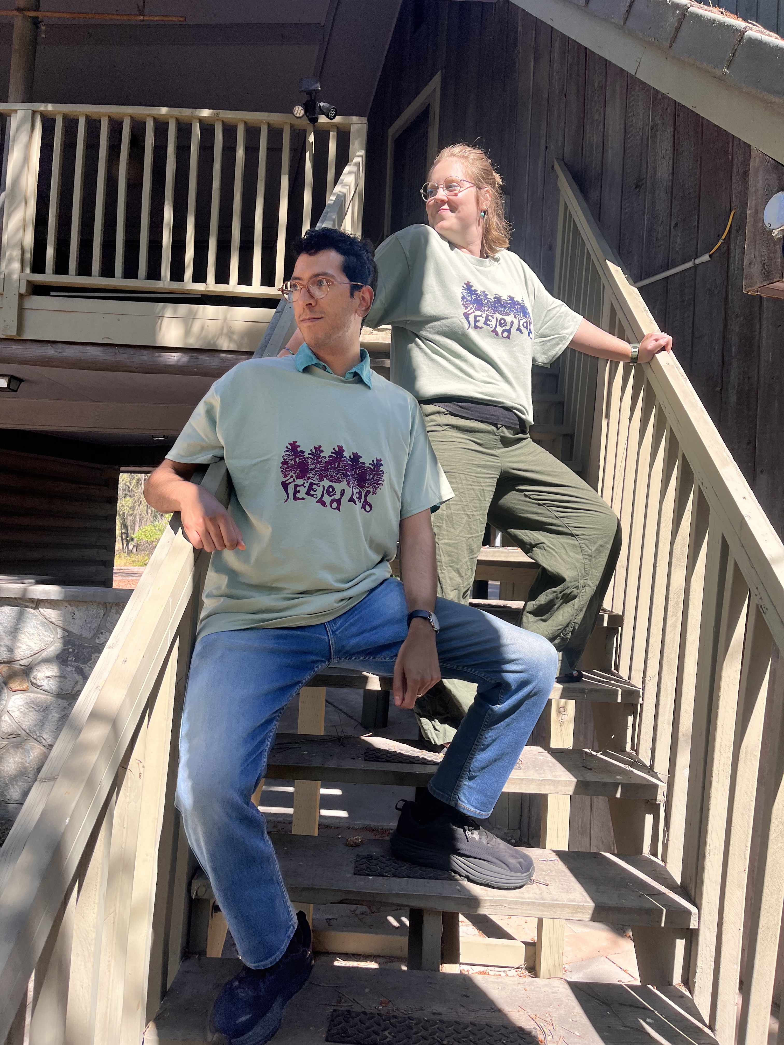 two people standing on the FEELed Lab stairs posing with the new FEELed Lab t-shirts. The shirts are light green with dark red and blue print