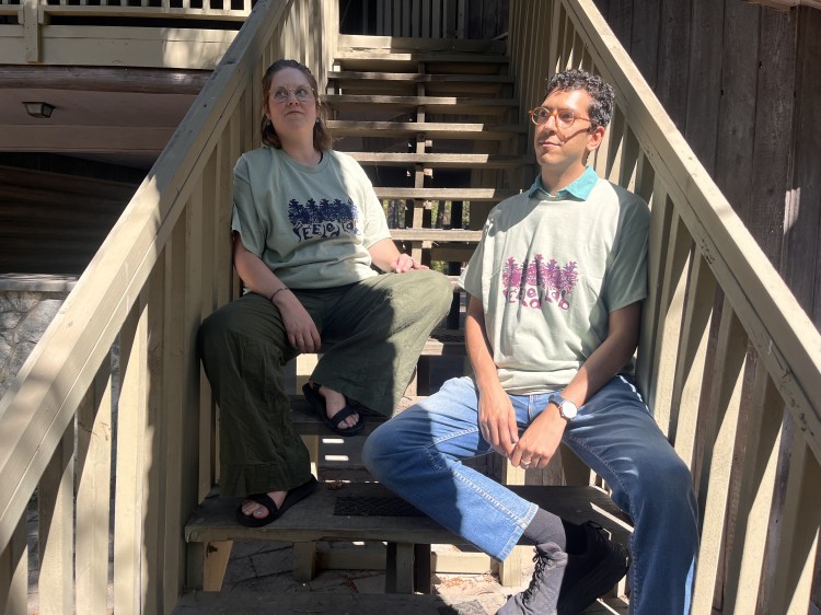 two people sitting on the FEELed Lab stairs in the sunshine wearing moss-green t-shirts with a printed FEELed Lab logo. They are posing catalogue-style