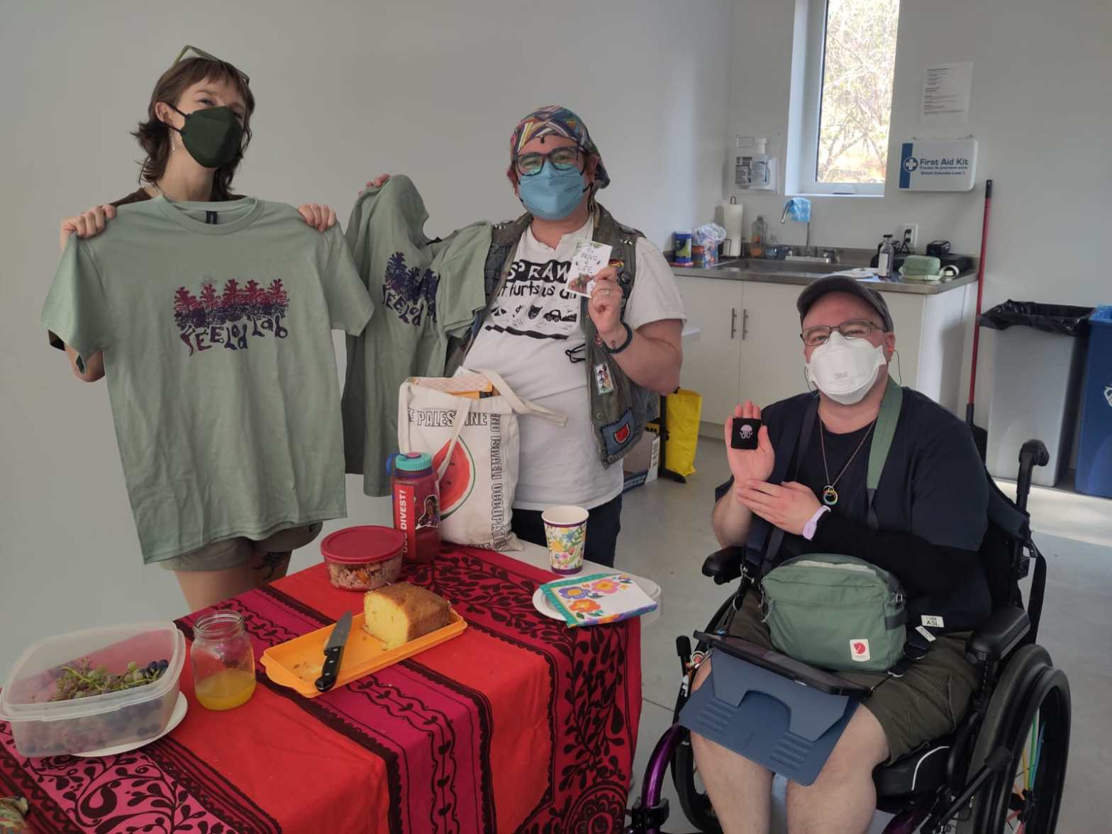 Three people holding up their crafty treasures including zines, a crossstitch jellyfish and the new FEELed Lab t-shirts at a table with a red tablecloth and cake inside woodhaven studio
