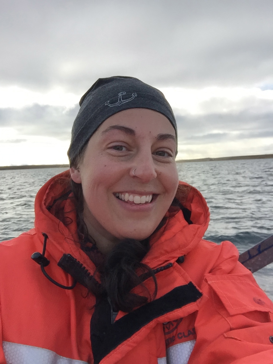Headshot of Sam on a boat. She is wearing an orange bright jacket and is smiling into the camera in front of a bright horizon