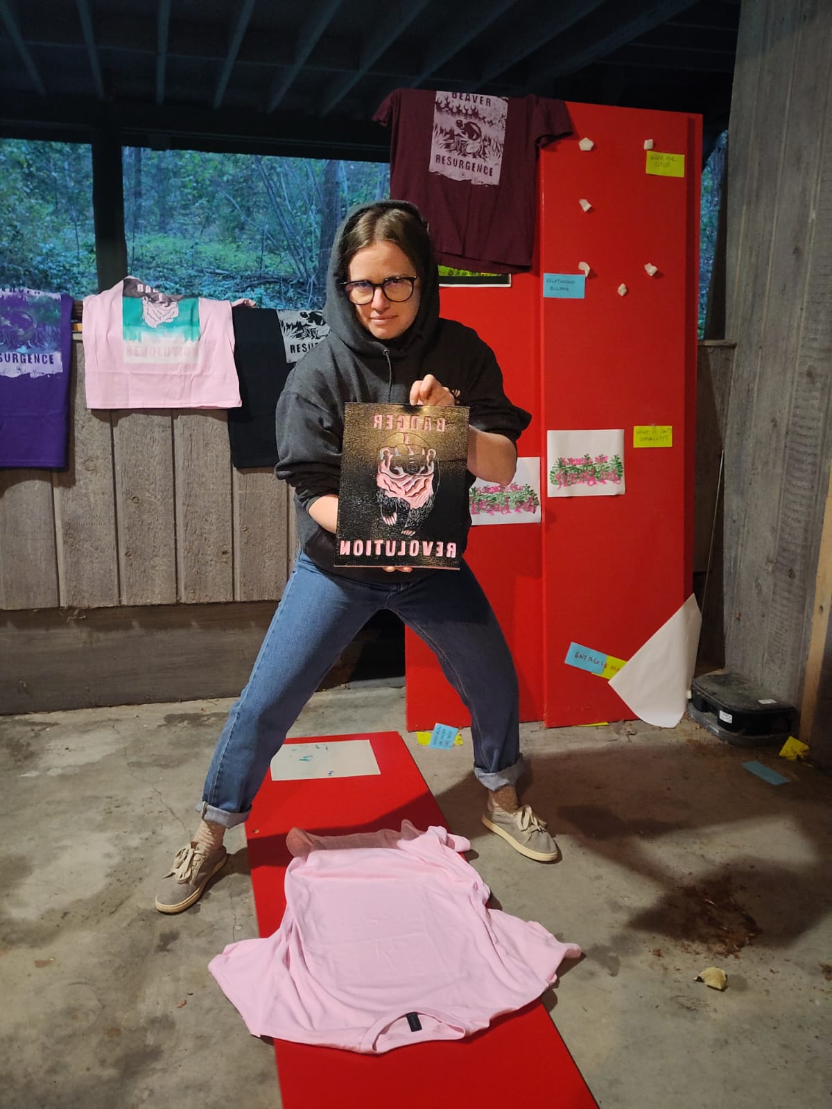 Jamie standing over a pink shirt with the ready-to-print linocut of the "Badger Revolution" in her hands. She is wearing a dark hoodie and is looking menacingly in the camera. There are many colorful printed shirts in the background