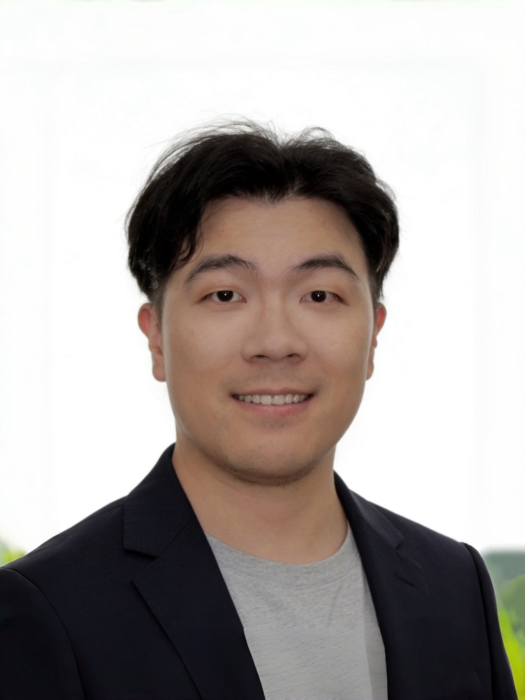 Headshot of Ming wearing a black blazer and a gray shirt in front of a green field and bright white background.