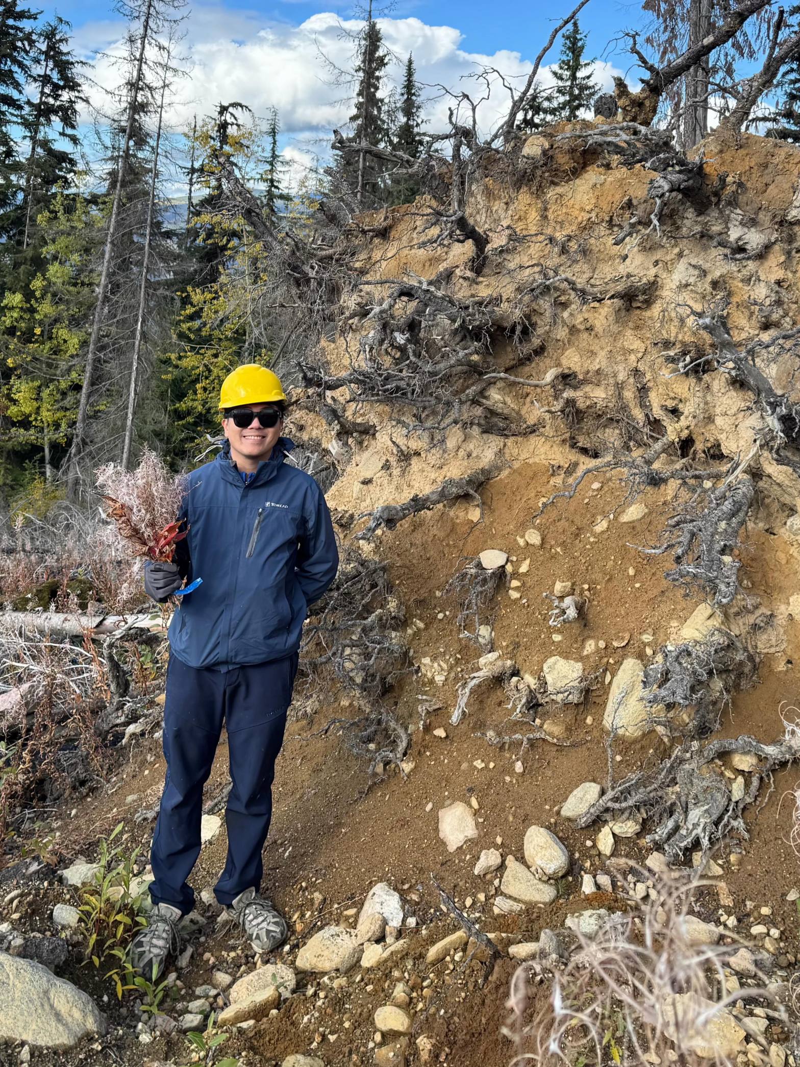 Full body shot of Ming standing next to a pile of dirt on the edge of a forest. He is wearing a bright yellow hardhat and there is a big forest in the background.