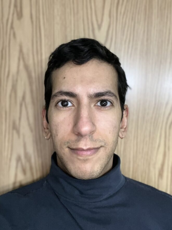 Headshot of Tareq wearing a black turtleneck sweater. He is standing in front of a bright wooden wall.