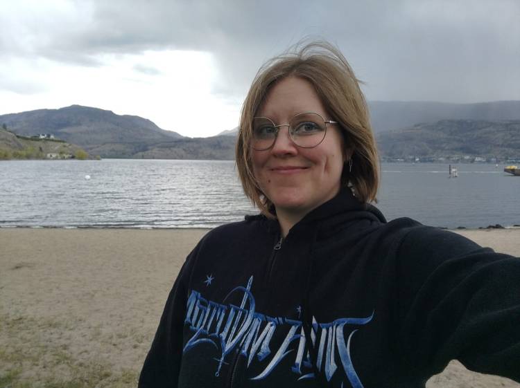Headshot of Julia in front of Okanagan Lake on a stormy day. Dey are wearing a black sweater with light blue writing